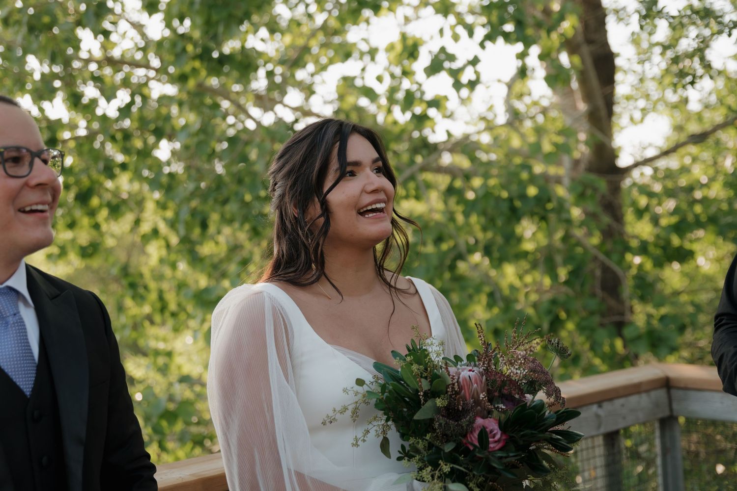 Wedding party celebrates outdoors with laughter and bouquets against a backdrop of trees and sunlight.