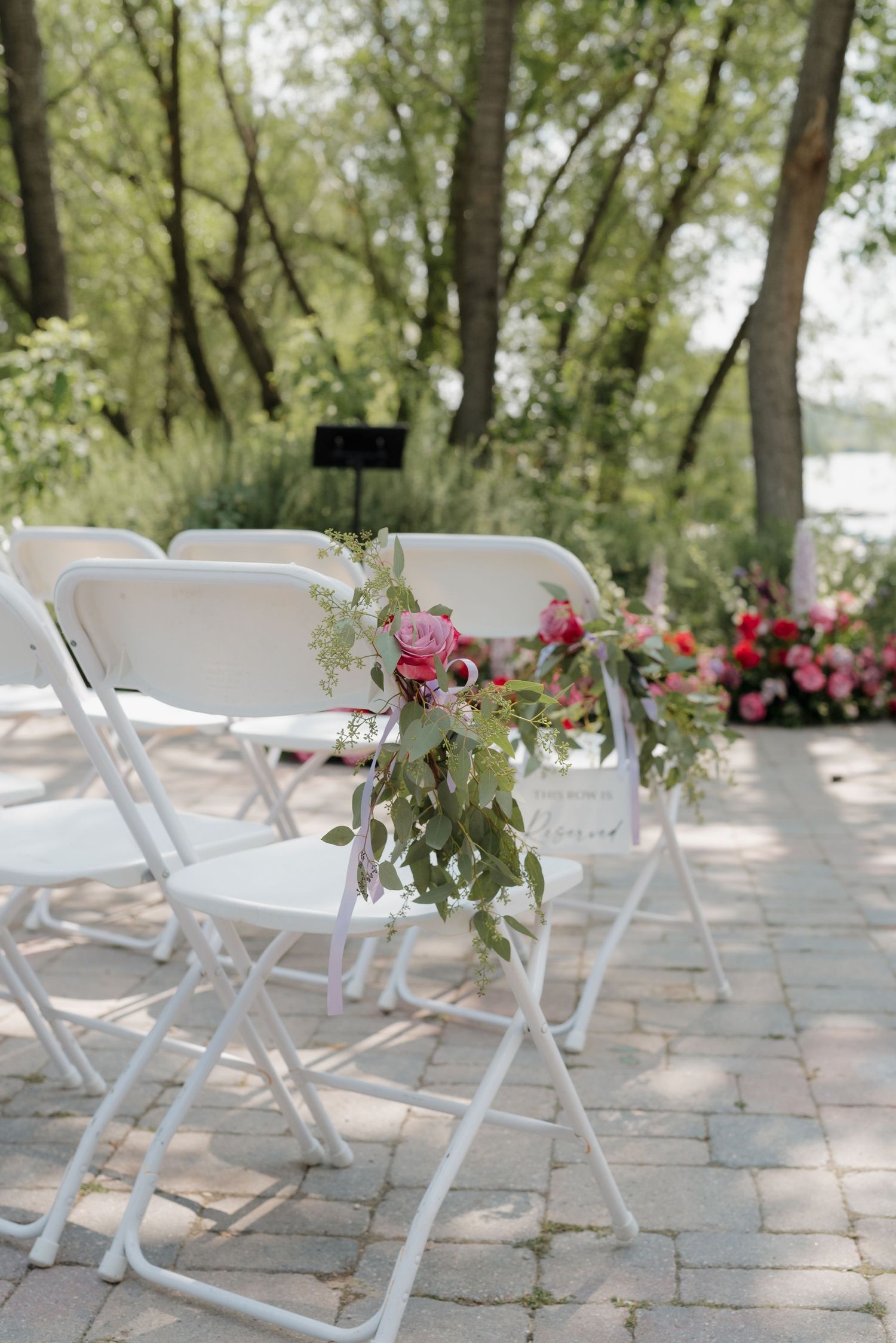 White folding chairs arranged on a stone patio with pink roses and greenery for an outdoor wedding ceremony.