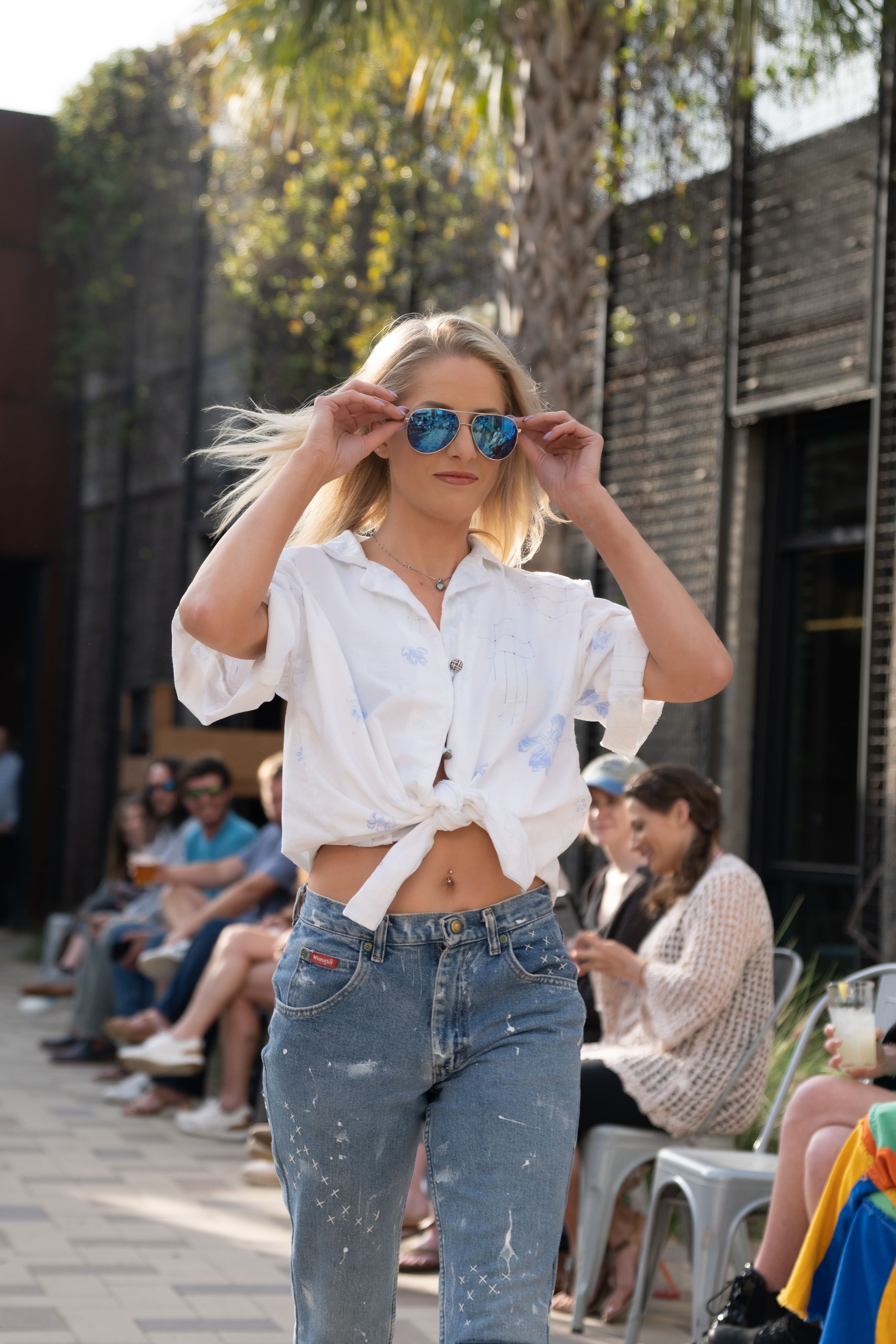 Woman in white tied shirt and distressed jeans poses outdoors on sunny day with blue sunglasses.