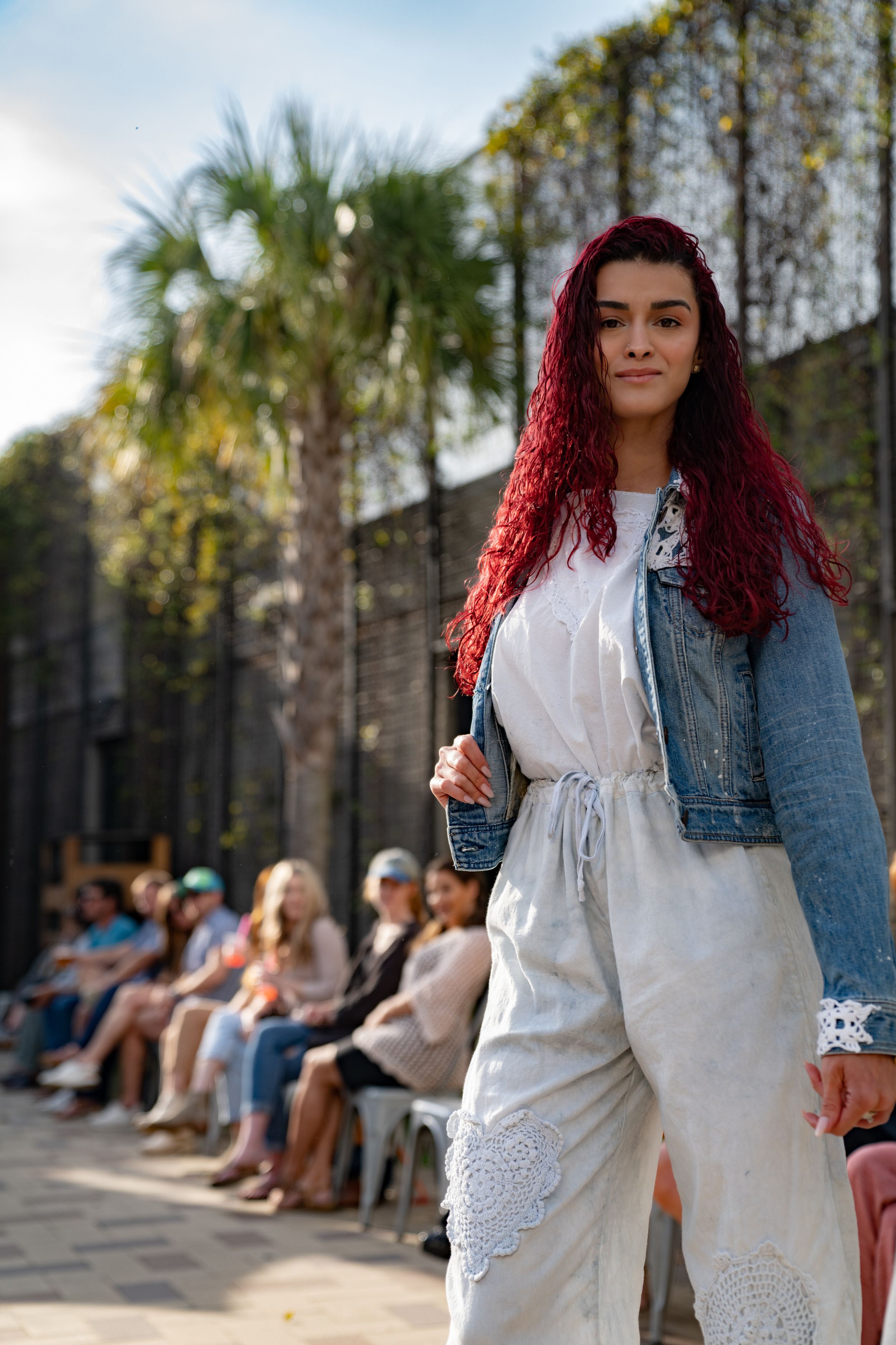 A fashion model walks down an outdoor runway wearing a white dress with lace details and a denim jacket.