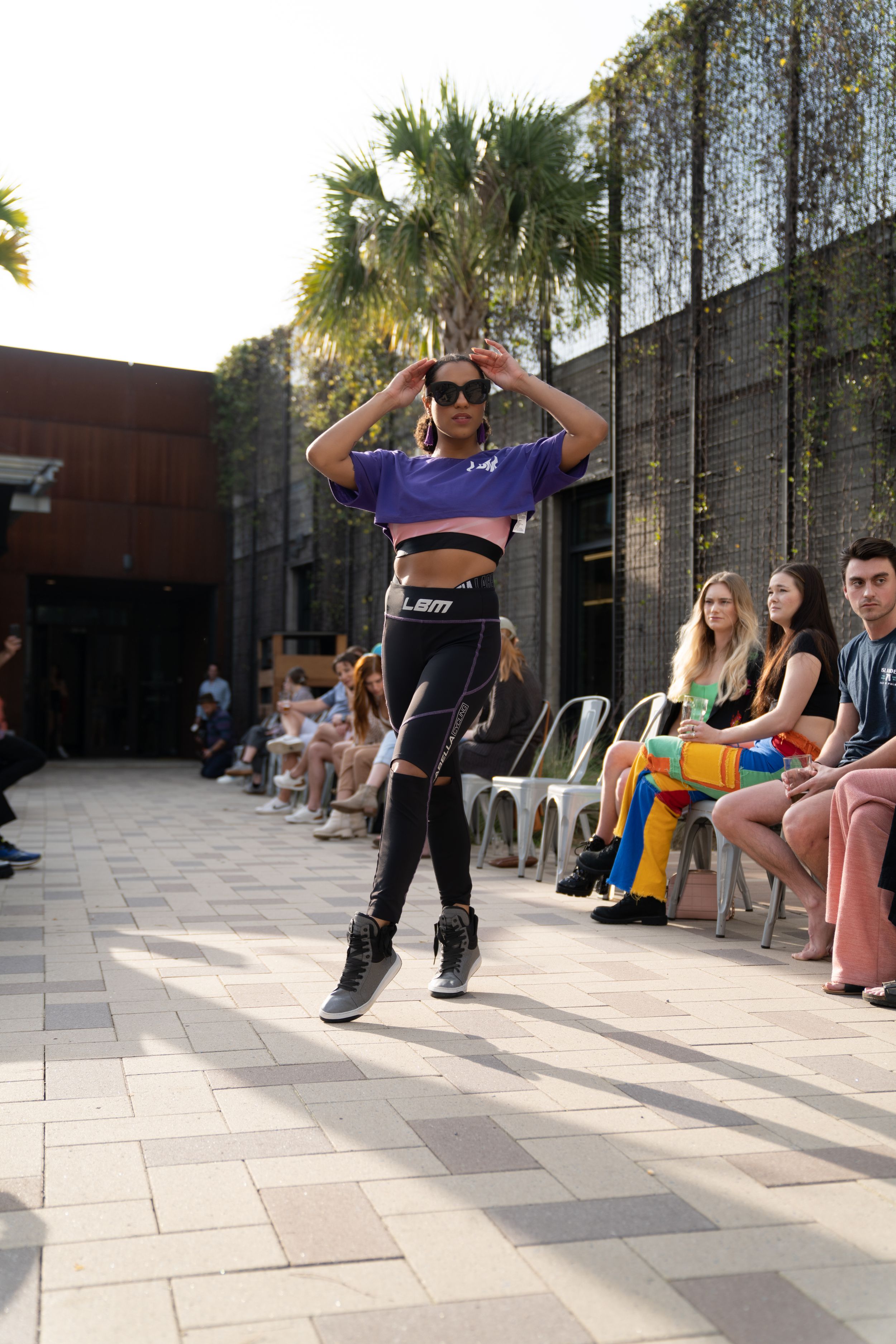 Person in athletic wear strikes dynamic dance pose on outdoor patio with audience in background.