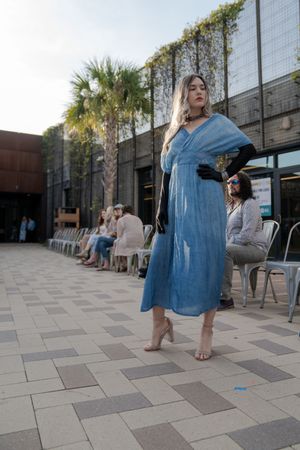 A person in a flowing blue denim dress poses on a paved outdoor area with palm trees in the background.