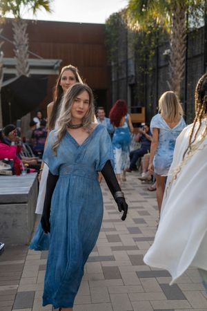 Models walk on an outdoor runway wearing blue and white dresses during a fashion show.