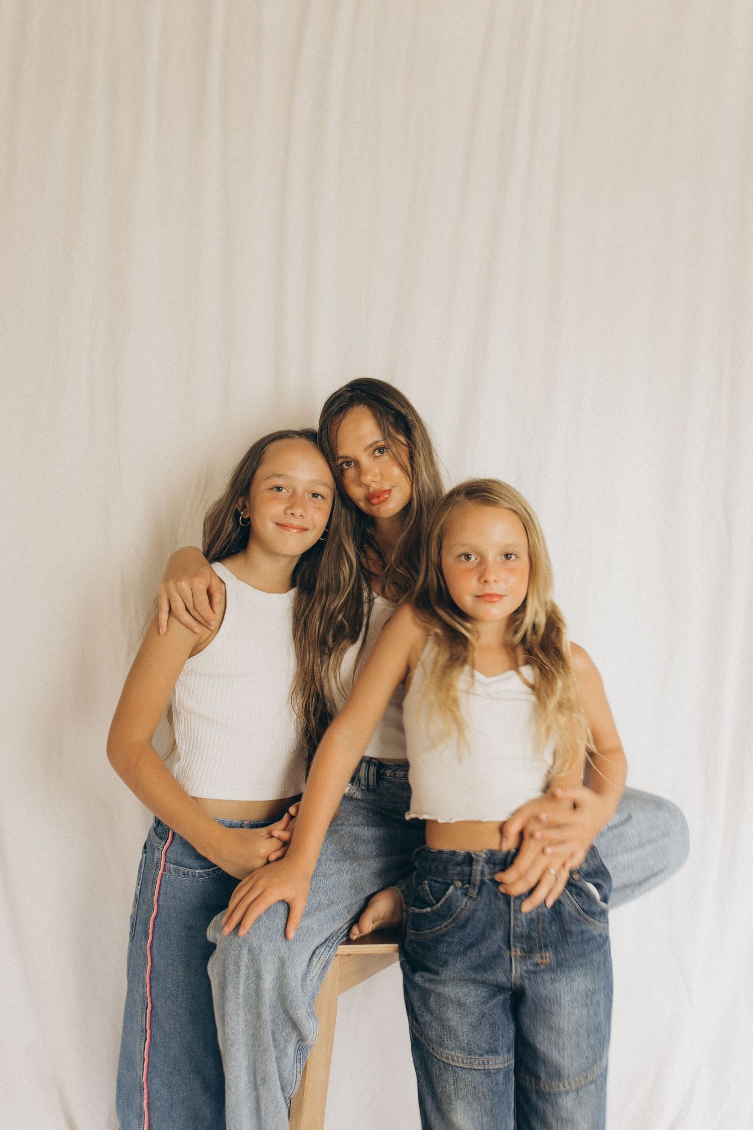 Mother in white top sits on stool embracing two daughters in white crop tops and jeans against white backdrop in New Zealand.