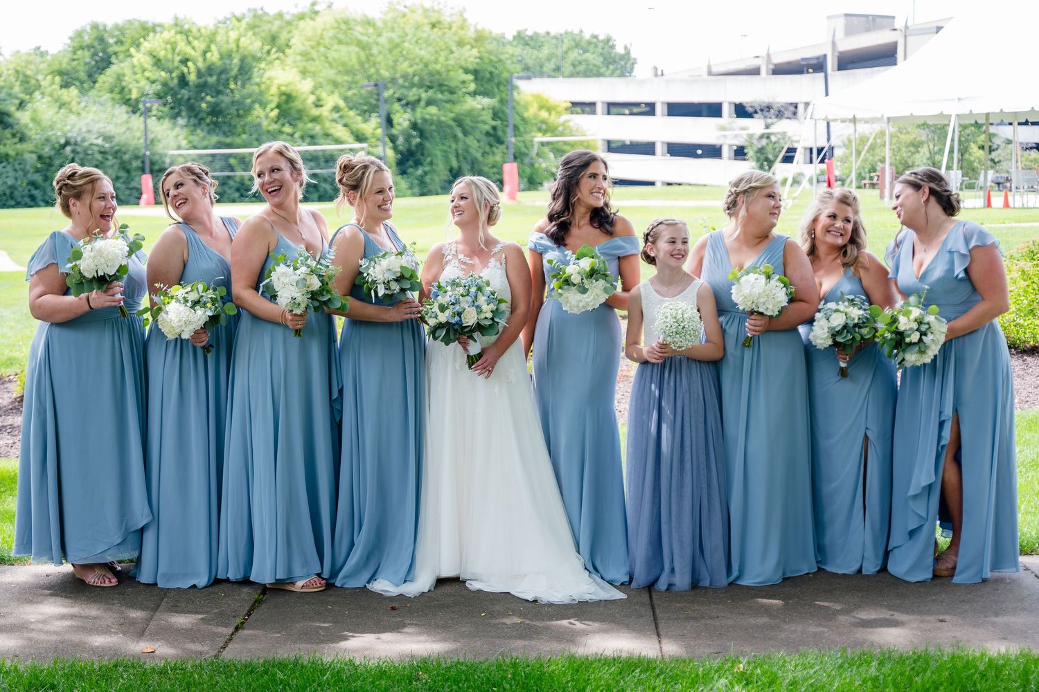 Wedding party in dusty blue bridesmaid dresses poses together outdoors on a summer day.