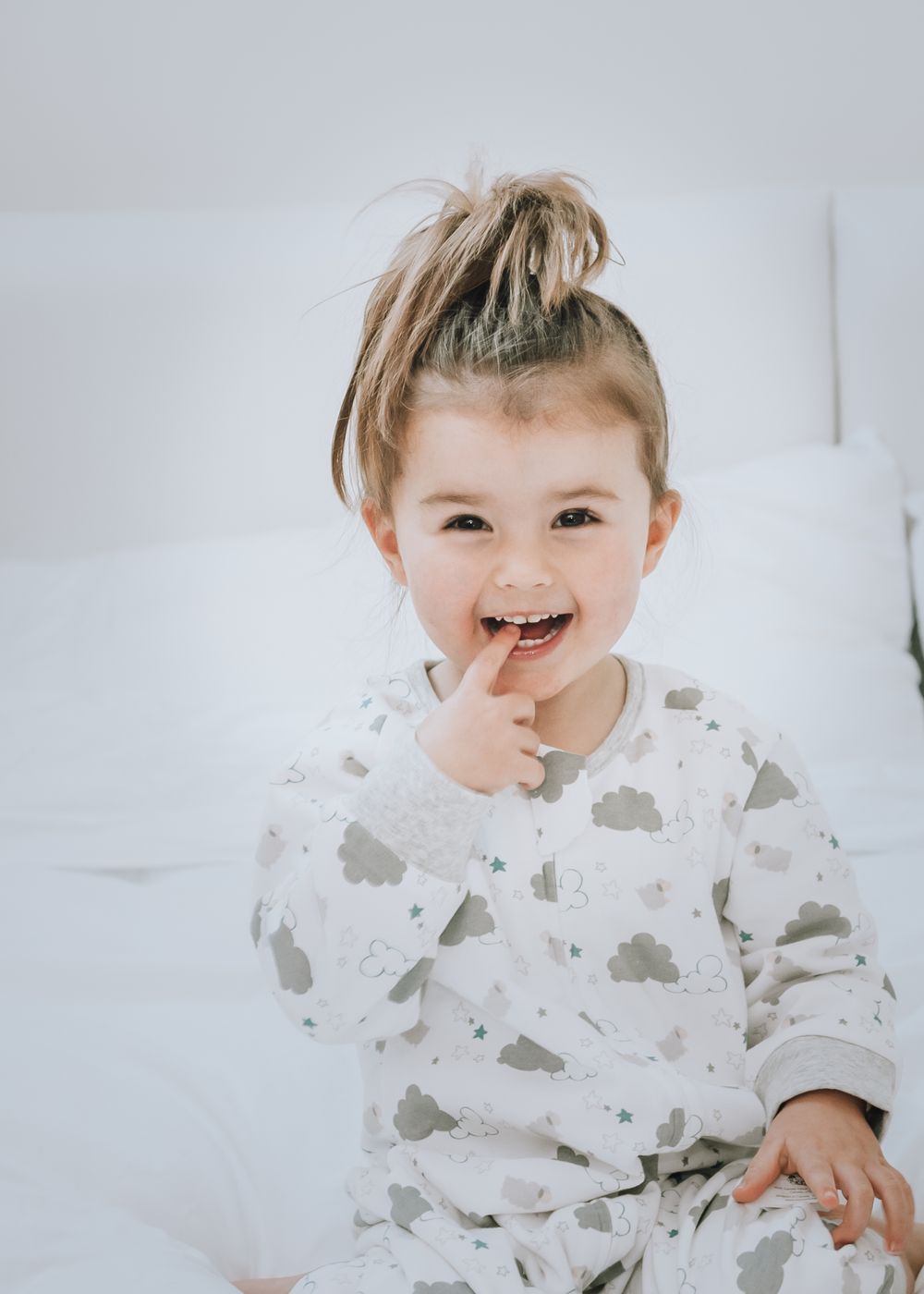 A small child in a cloud-patterned sleepsuit plays and laughs on a white bed.
