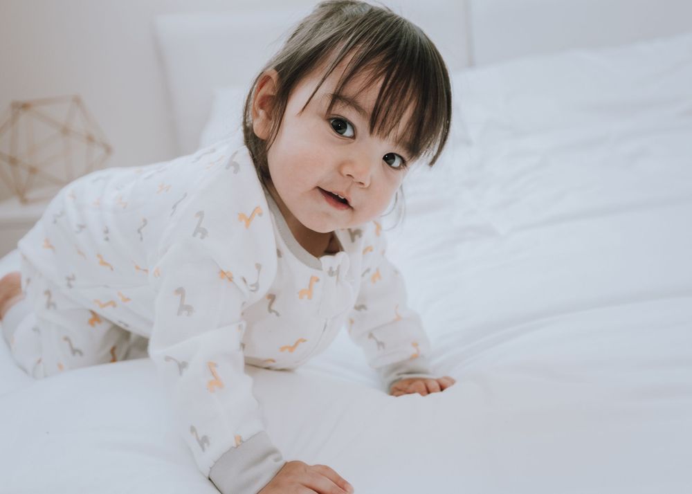 A young child in a white dress lies on white bedding while looking at the camera with a sweet expression.