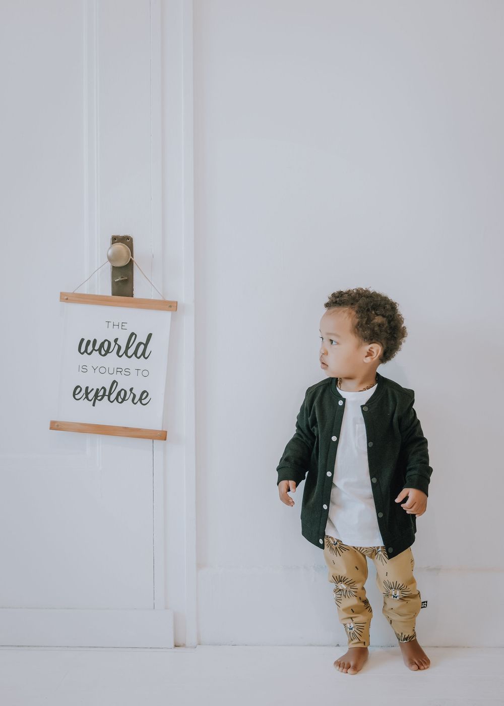 Small child in black jacket and khaki pants stands against white wall with minimalist wall art.