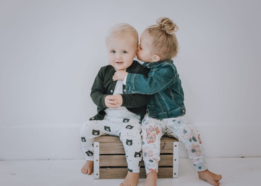 Two young children sit together on a wooden bench wearing coordinating outfits in white and green tones.