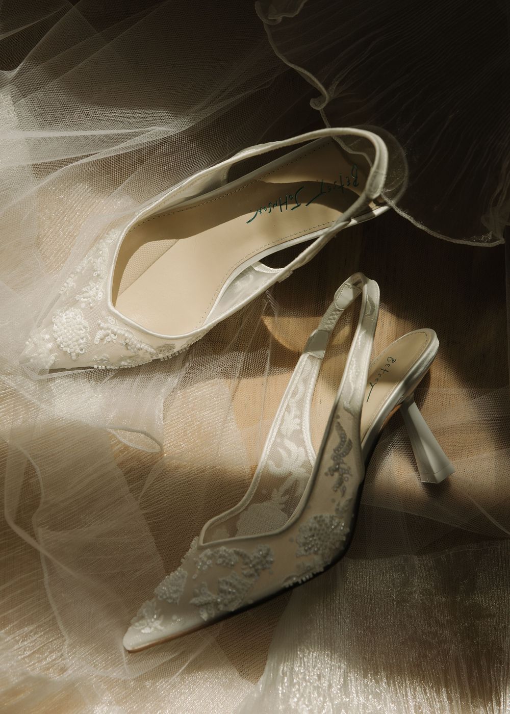 Delicate white bridal shoes with lace detailing photographed in dramatic lighting.