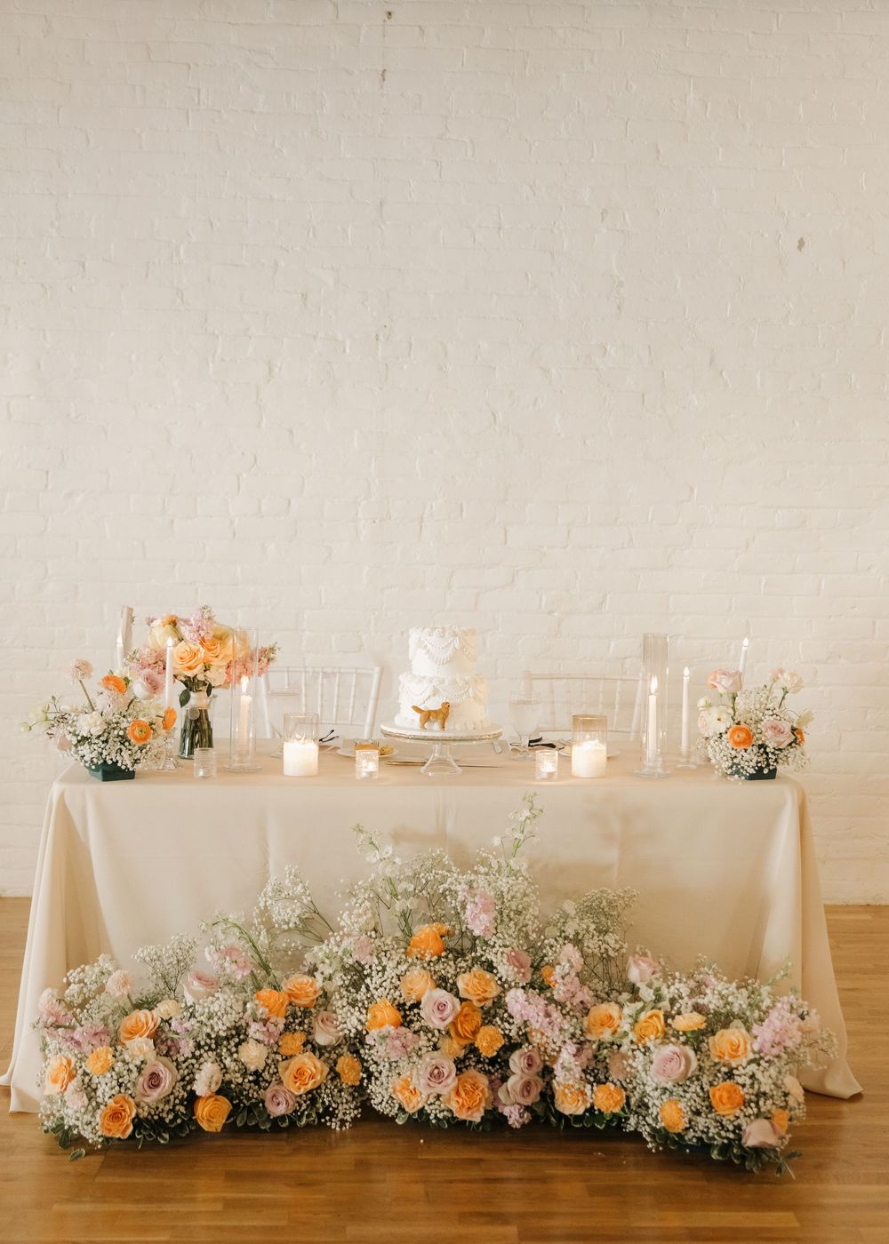 Elegant wedding reception table with peach and white floral arrangements and candles against white wall.
