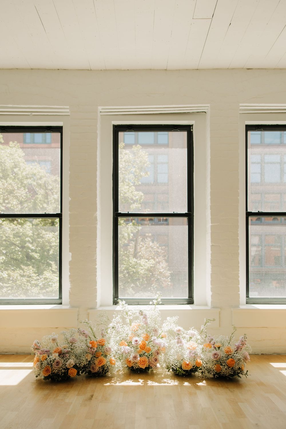 Three large windows with delicate floral arrangements placed beneath on hardwood floors.