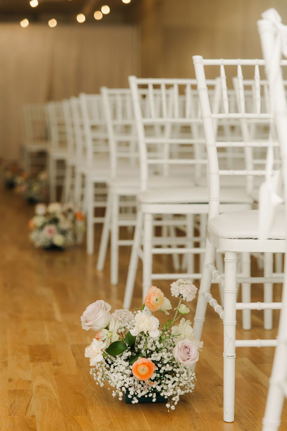 White wedding ceremony chairs lined up with delicate floral arrangements of roses and baby's breath on a wooden floor.