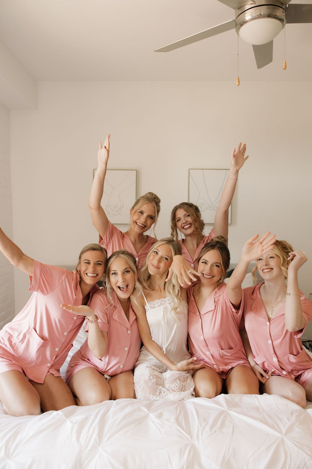 Group wearing matching pink robes celebrating joyfully on a white bed during wedding preparations.