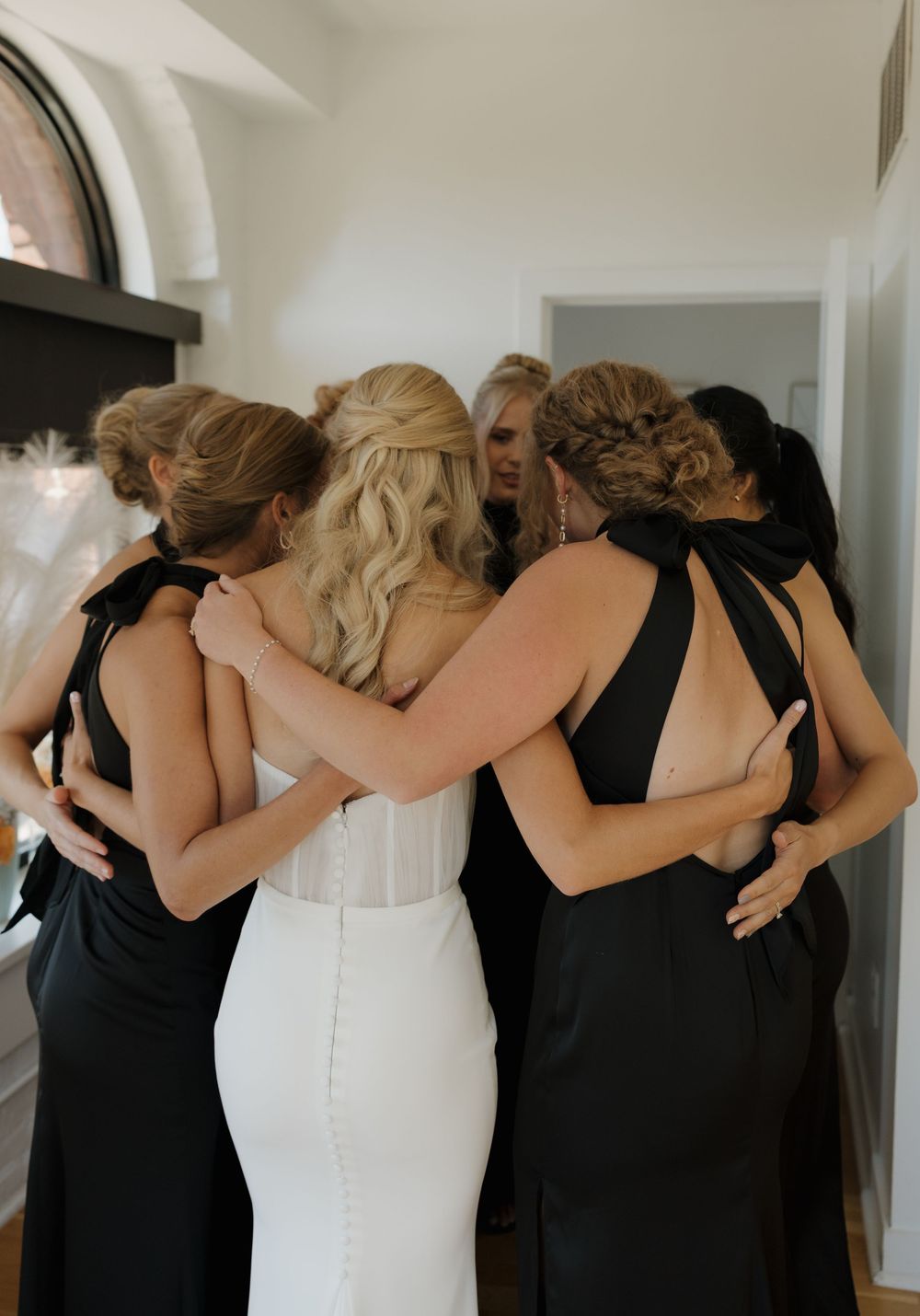 Bride in white dress and bridesmaids in black dresses embracing in a heartfelt group hug.