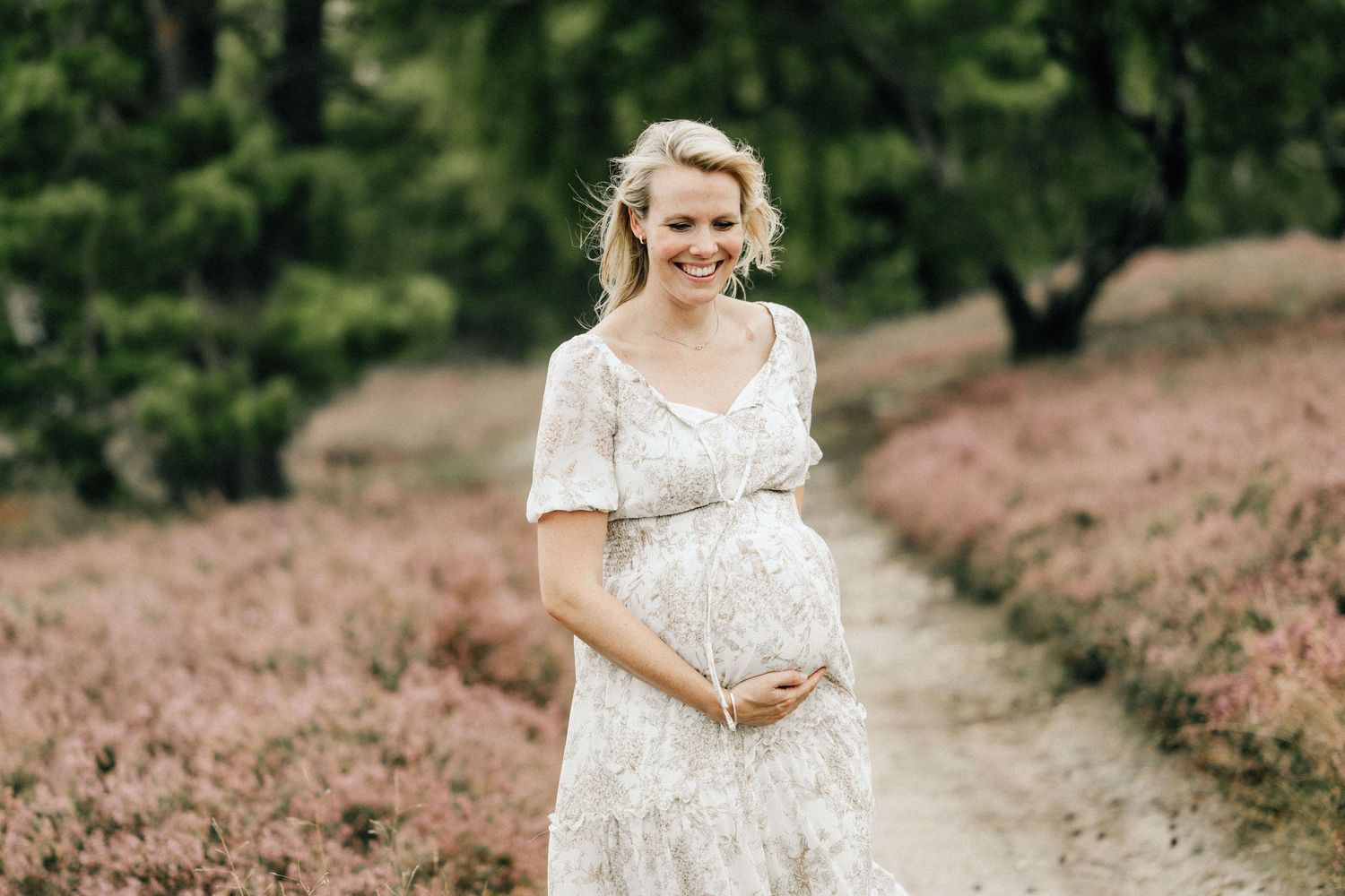 A pregnant woman in a white lace dress stands on a path surrounded by blooming pink heather flowers in nature.