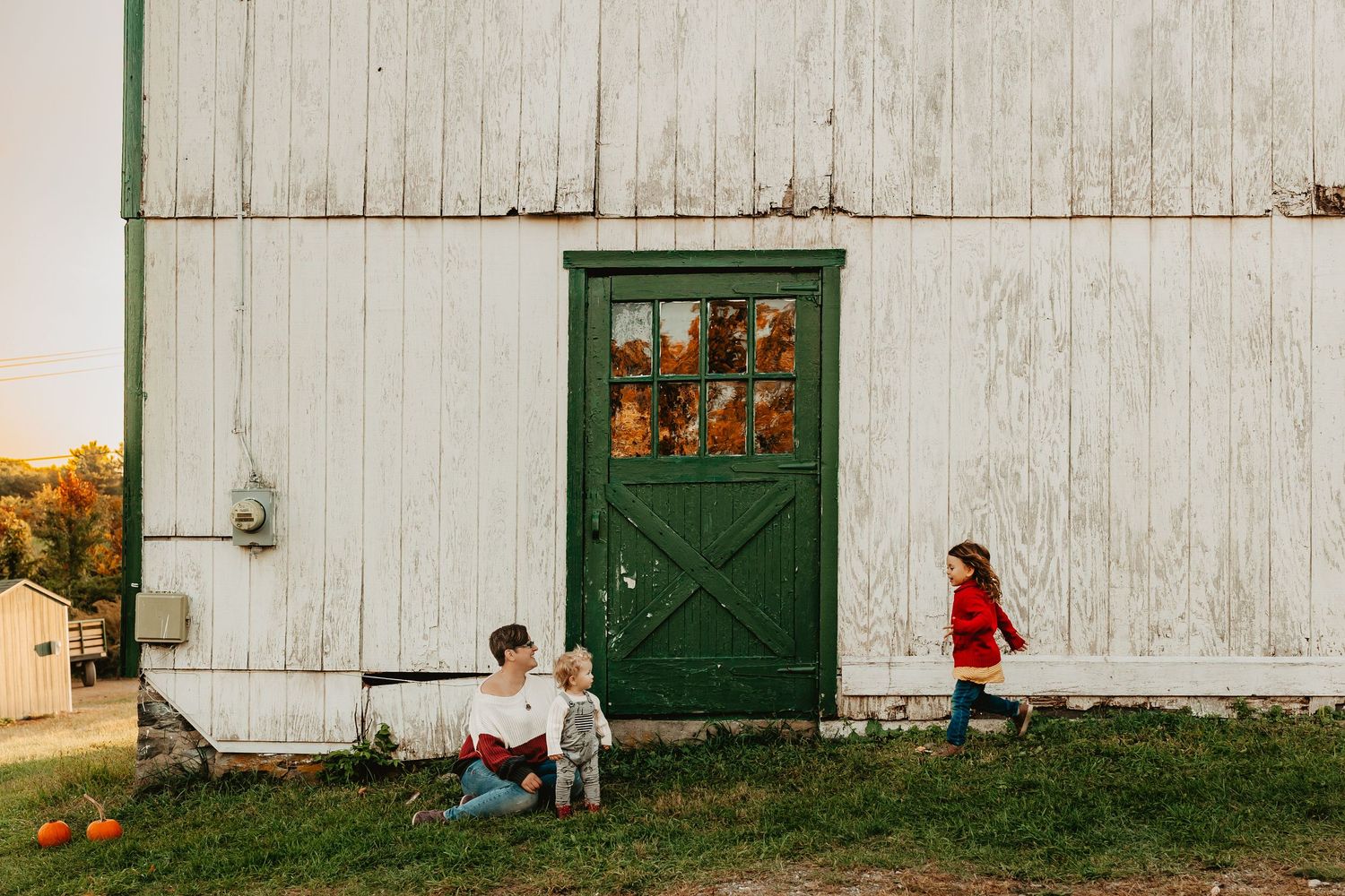 Children play near a rustic green barn door on a white wooden building at sunset.