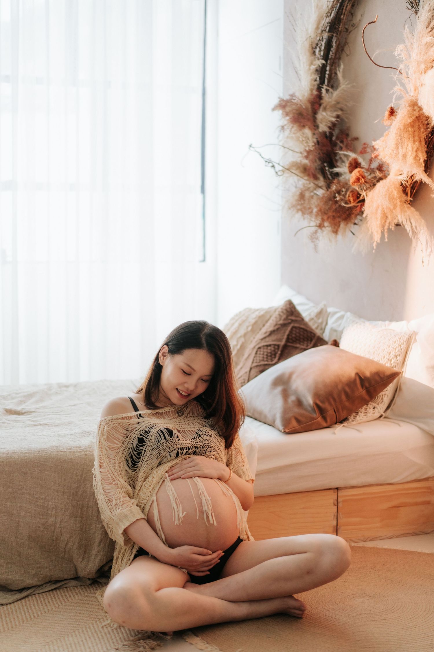 Expectant mother in cozy sweater sits on floor by window with natural light and dried floral decor.