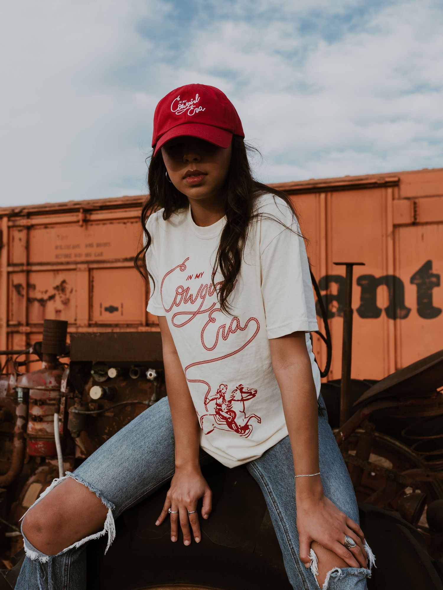 Young woman in a Cowgirl Era graphic tee and red cap posing near rustic freight train cars.