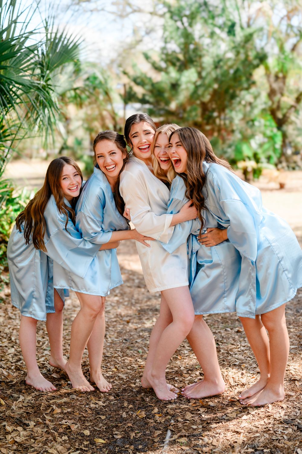 Bridesmaids in matching light blue robes share joyful moments together in an outdoor garden setting.