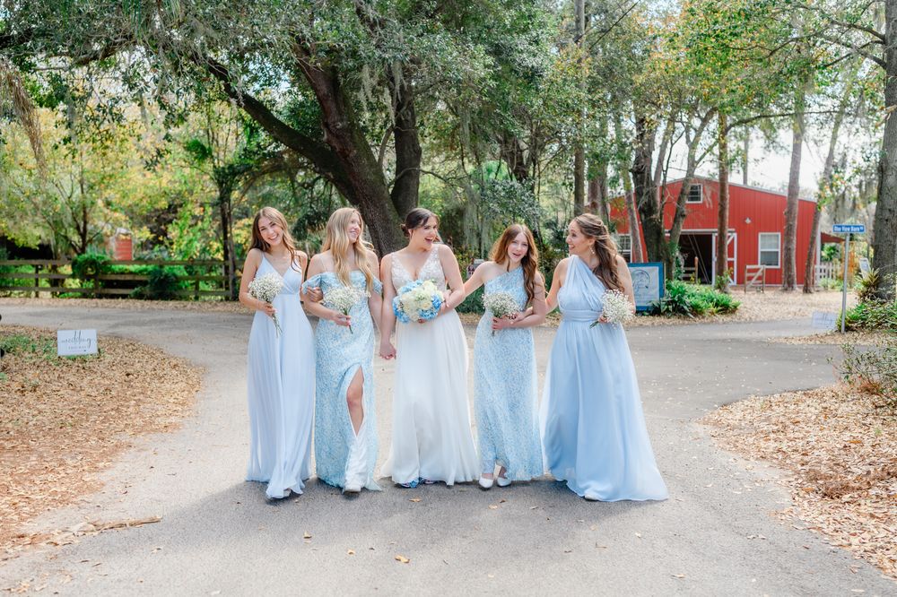 Wedding party in flowing light blue dresses walks together along a tree-lined pathway.