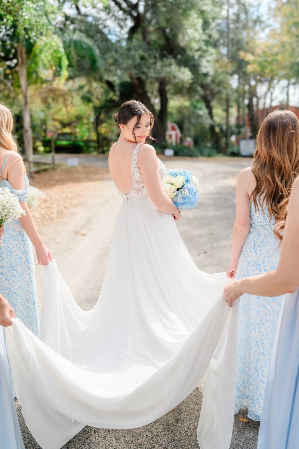 Bridesmaids in light blue dresses holding up the bride's wedding dress train outdoors.