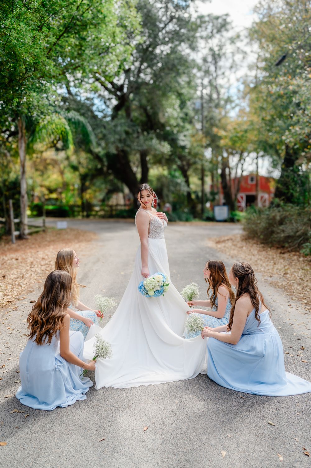 Bridesmaids in light blue dresses help arrange a bride's wedding gown train on a tree-lined path.