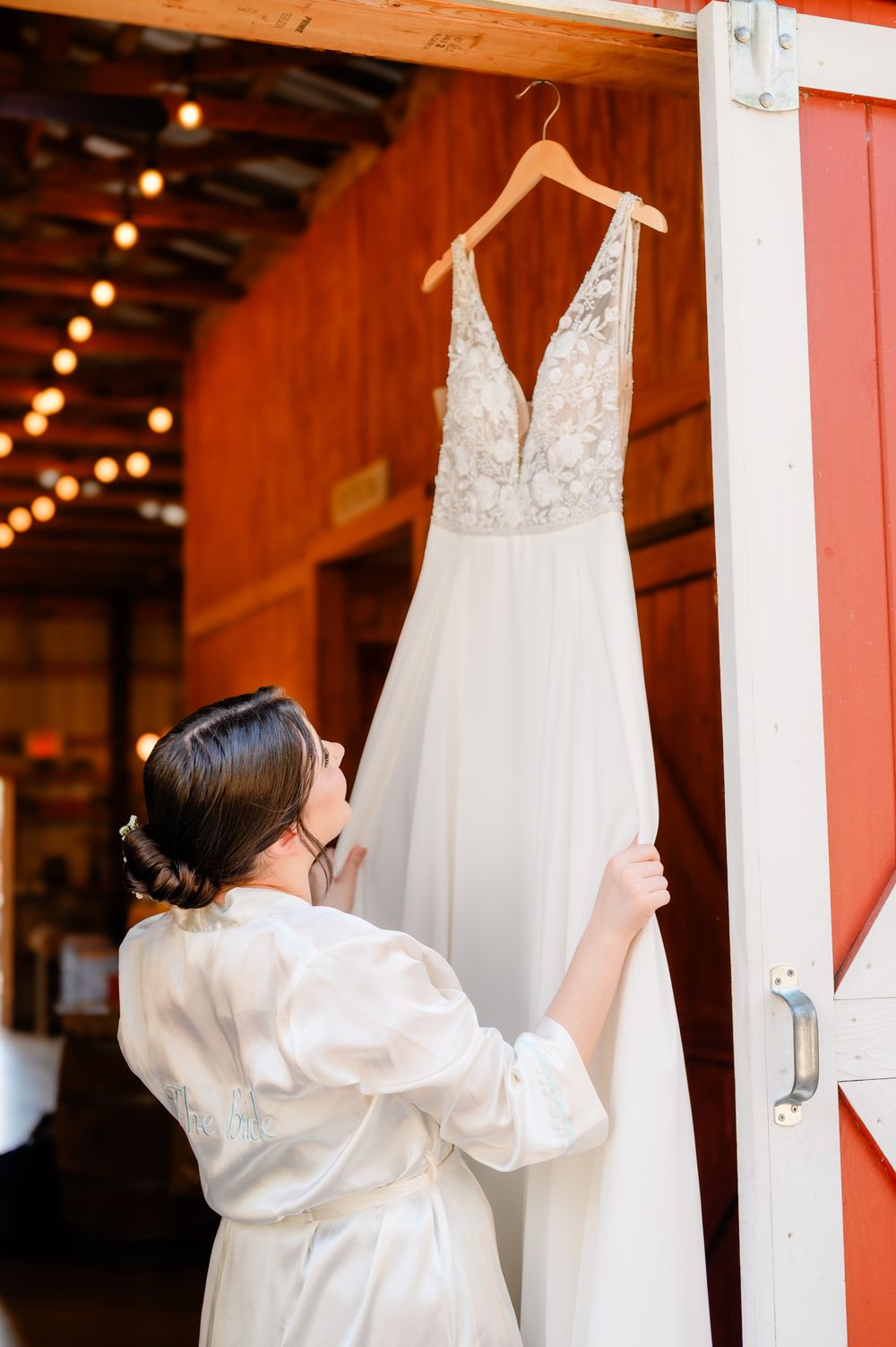 White wedding dress hanging in a rustic venue with string lights and wooden beams.