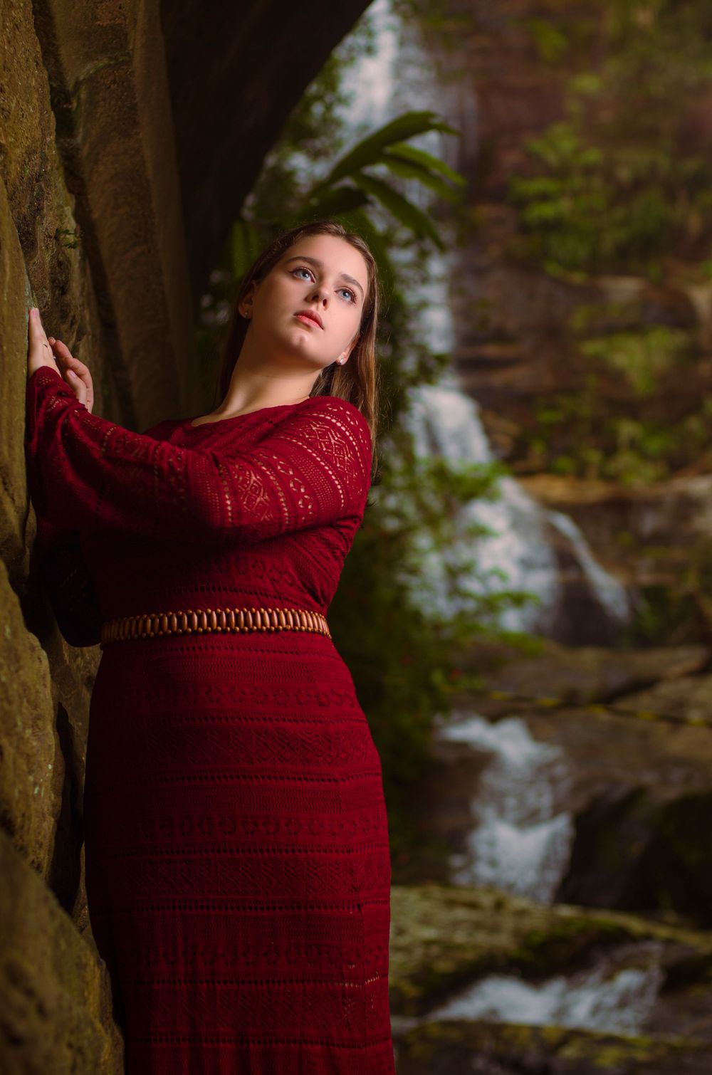 Ensaio feminino em ambiente natural. Mulher com vestido longo bordô posando em uma parede de pedra abaixo de uma ponte com cachoeira.