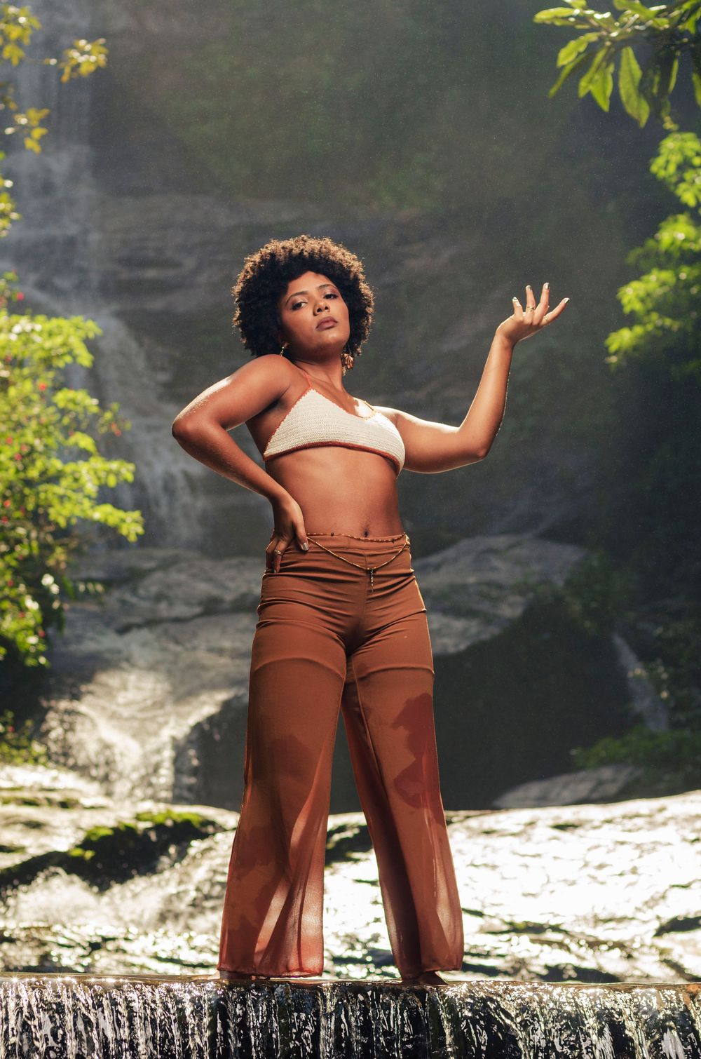 Ensaio de dança na natureza. Modelo negra com cabelo black power e pose artística, em frente à cachoeira com queda d'água.