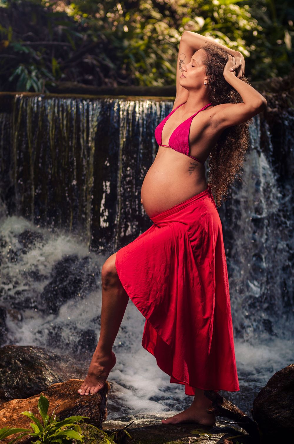 Ensaio de gestante na cachoeira. Mulher grávida com cabelo cacheado, com biquíni rosa e saia vermelha, posando em rochas.