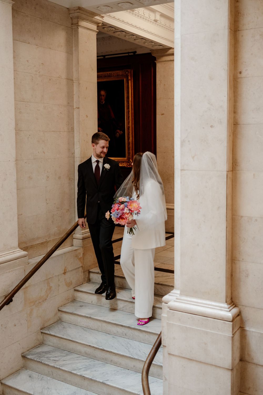 A couple in wedding attire stands on marble steps by elegant stone columns in a classic architectural setting.