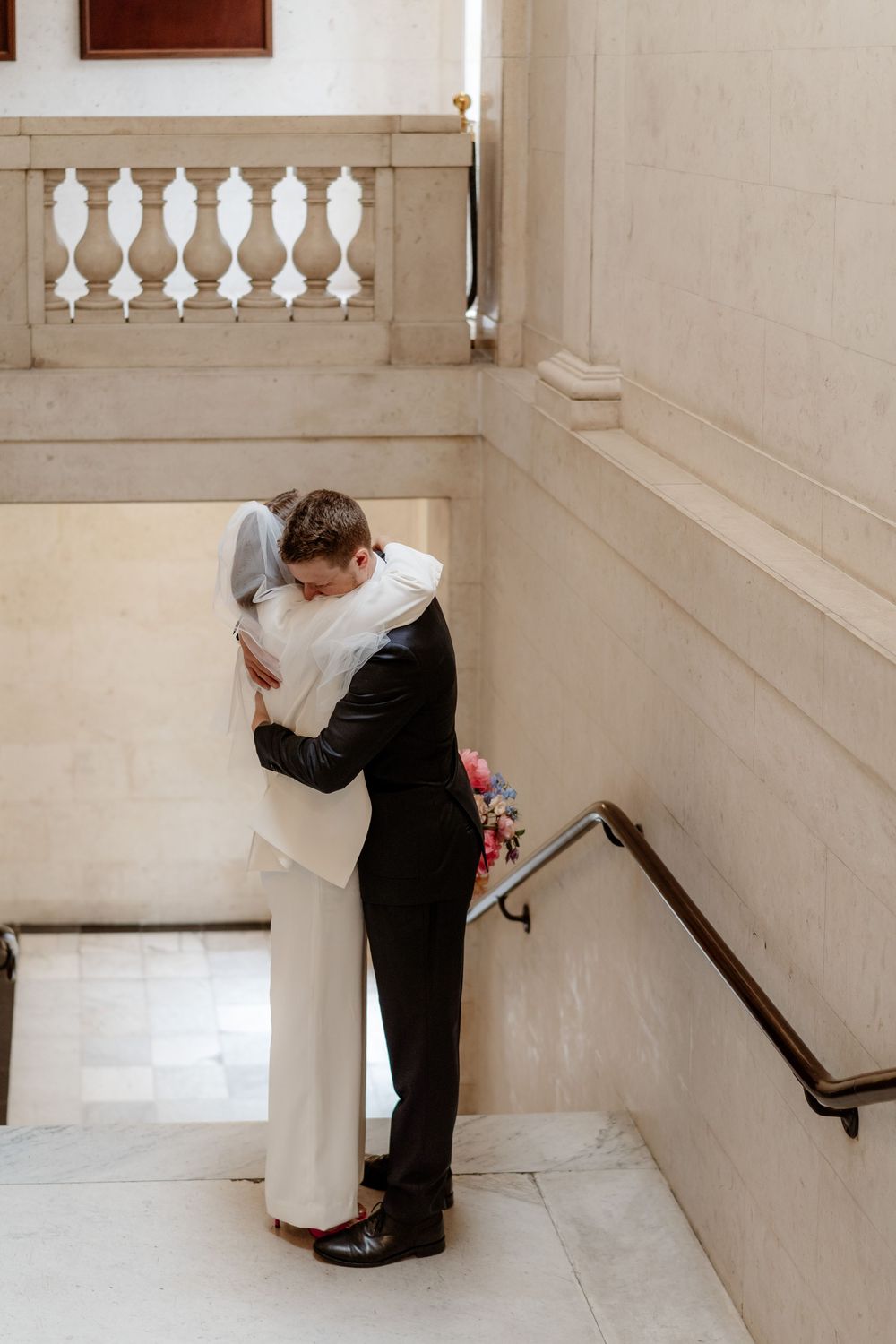 Two people share an intimate embrace on an elegant white marble staircase with decorative balusters.