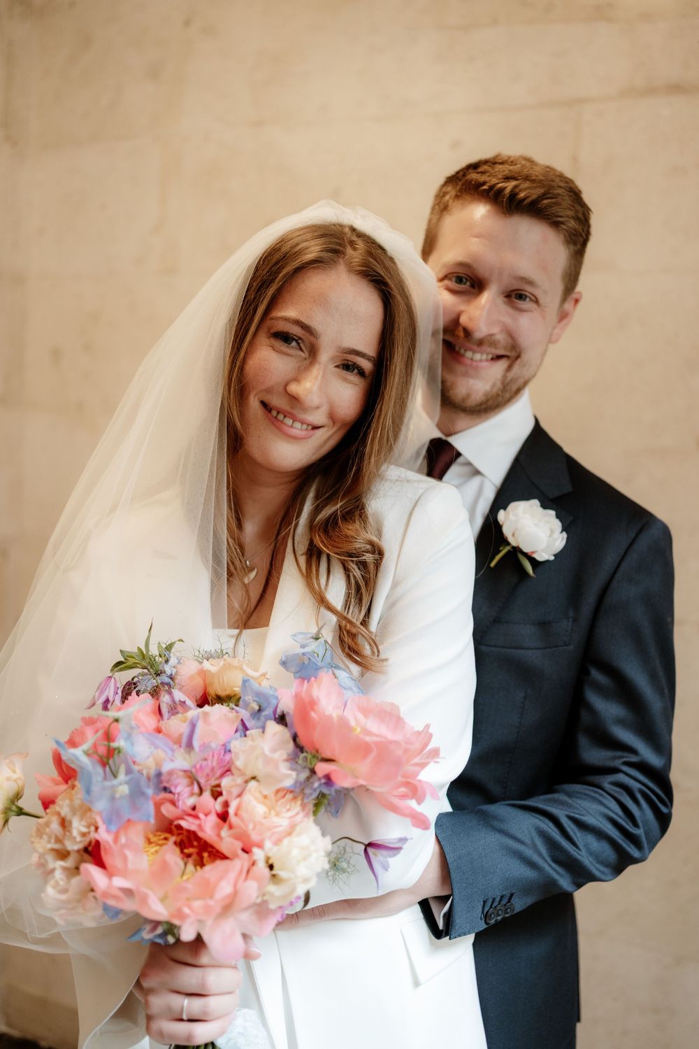 Romantic wedding couple poses together with a beautiful bouquet of pink and pastel flowers against neutral backdrop.