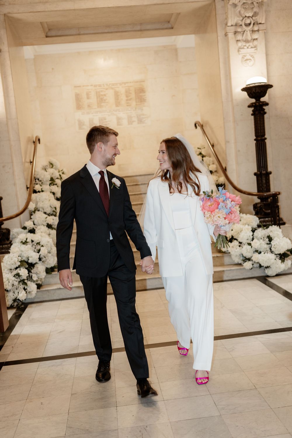 A couple in formal attire shares a playful moment in a grand marble hallway decorated with white floral arrangements.