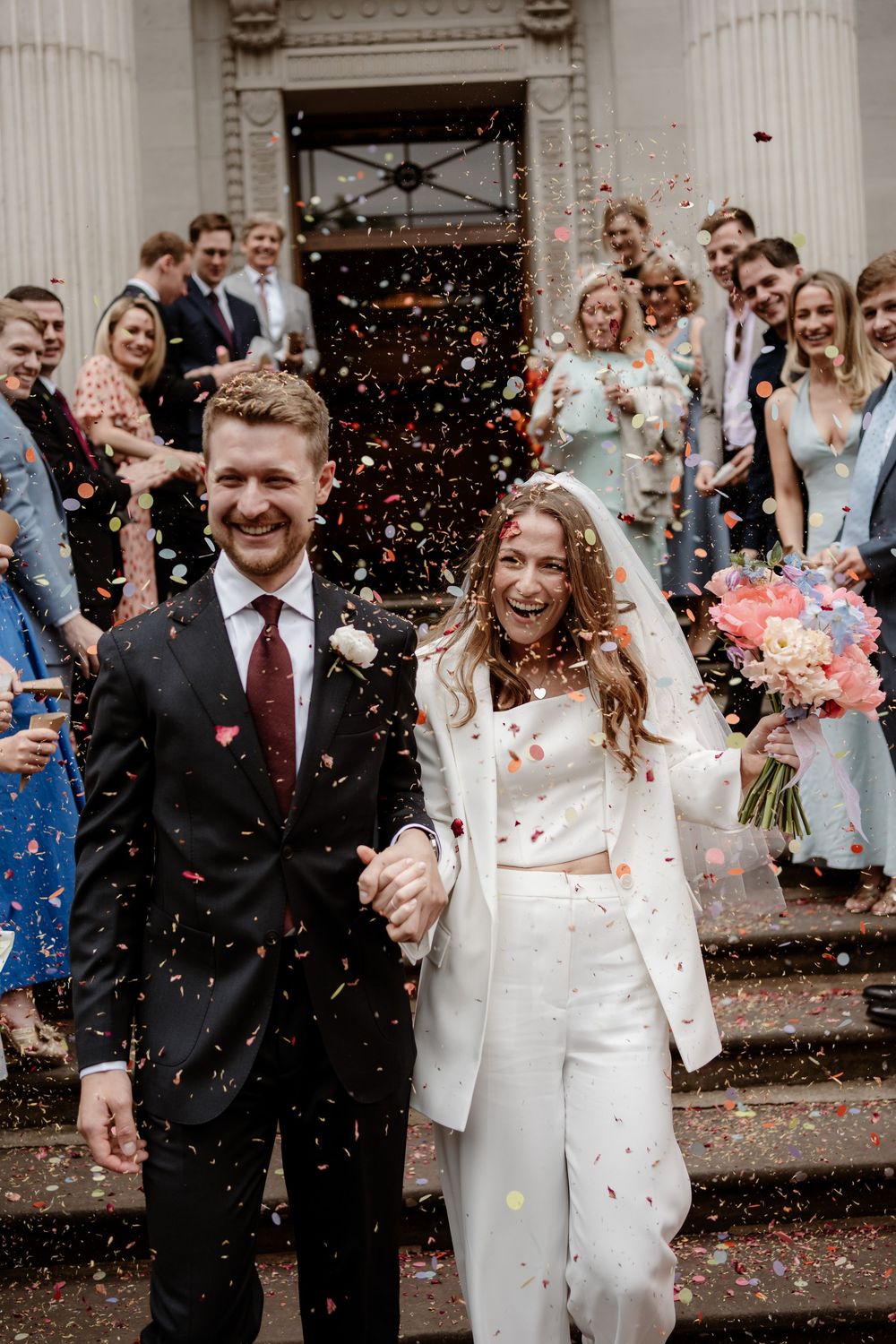 A joyful wedding couple exits their ceremony while guests shower them with colorful confetti on the steps.