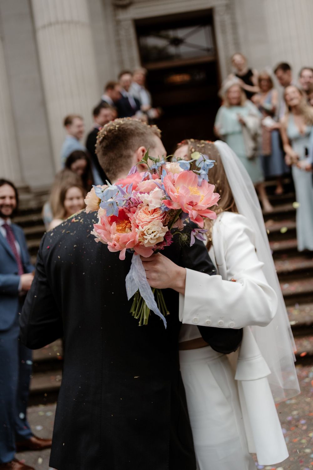 Newlyweds share romantic kisses behind pink and white bouquets on church steps while guests look on.