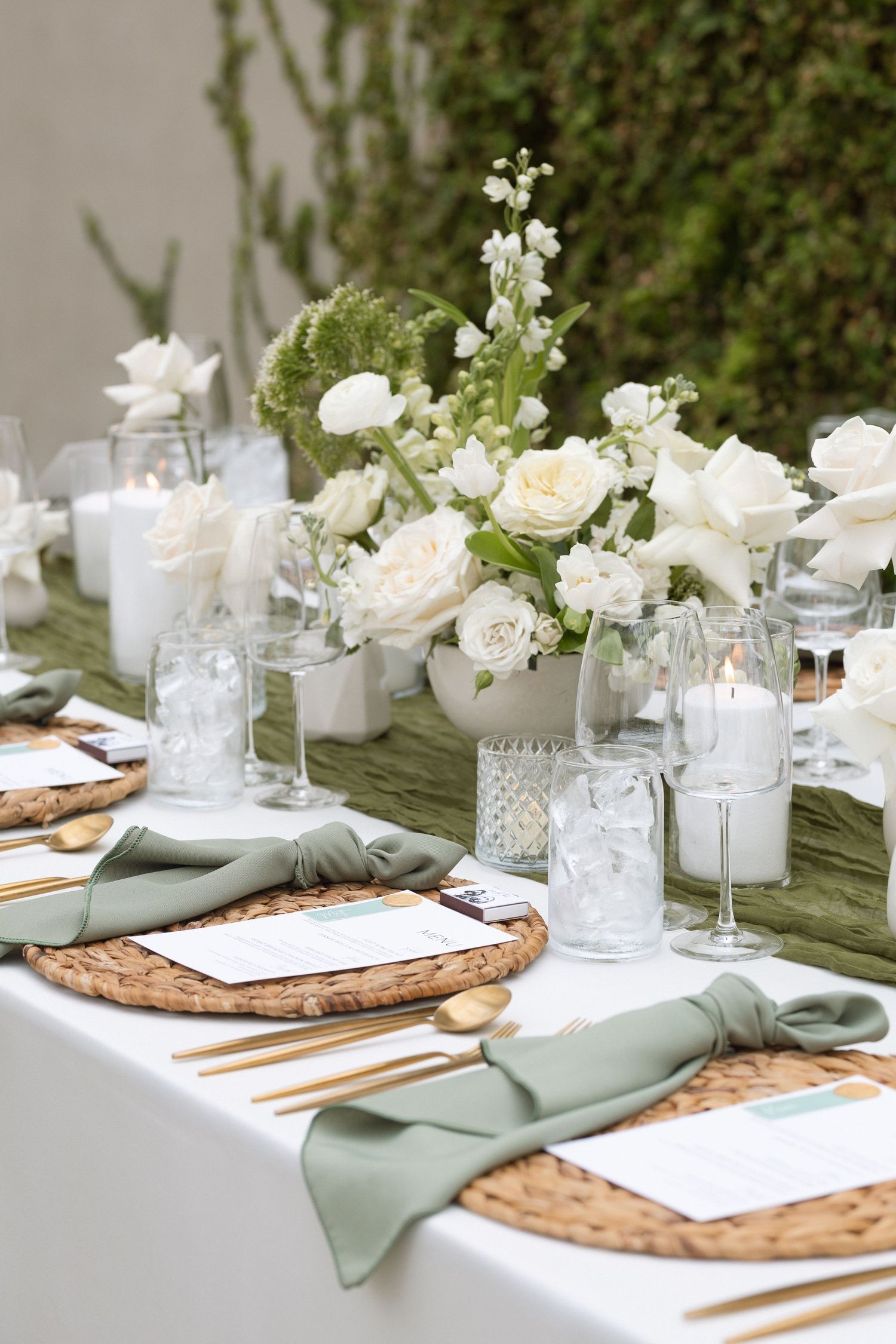 Elegant table setting with white flowers, sage green napkins, woven placemats and glass candle holders in natural light.