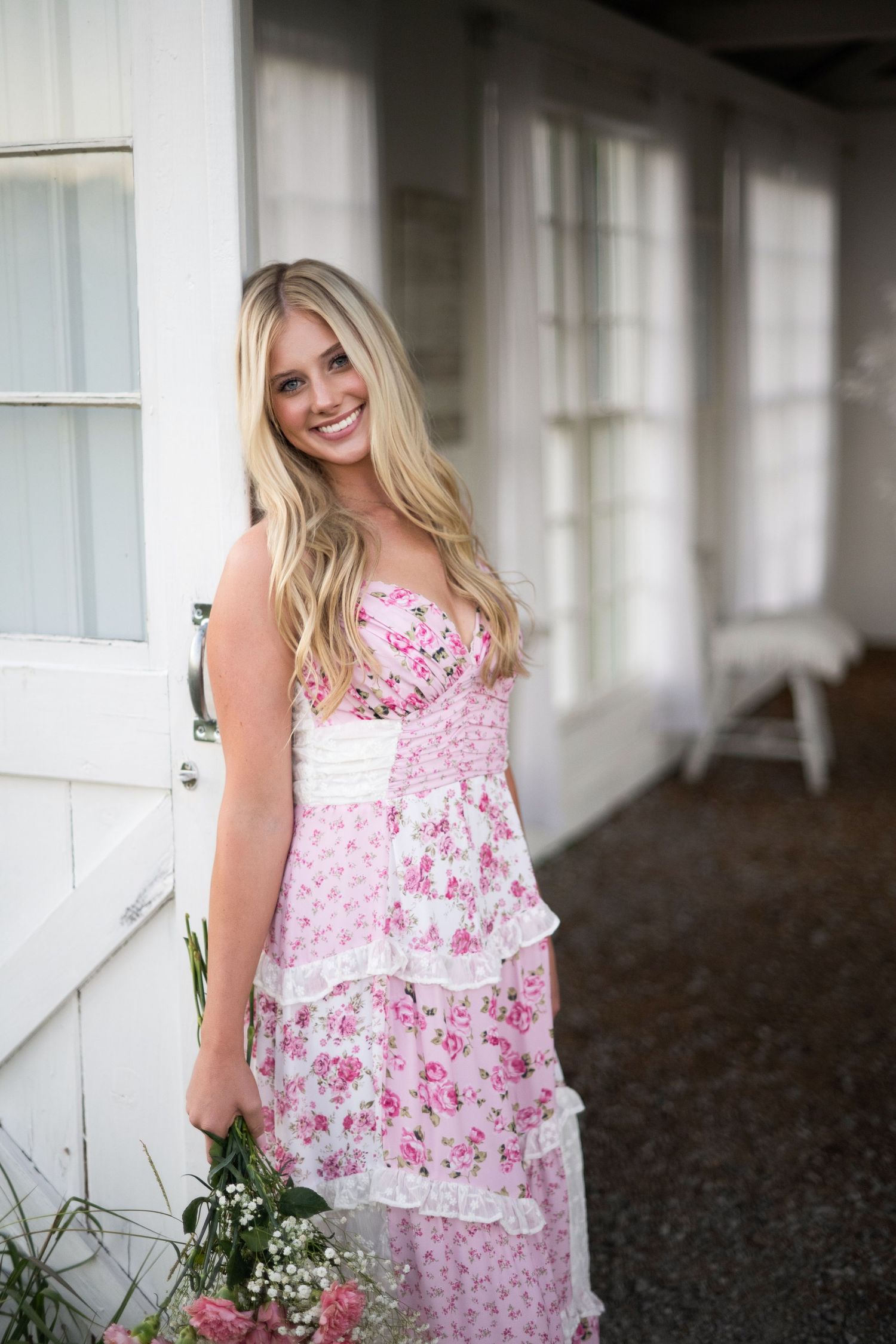 Woman in pink floral sleeveless dress poses in front of white building in outdoor portrait series.