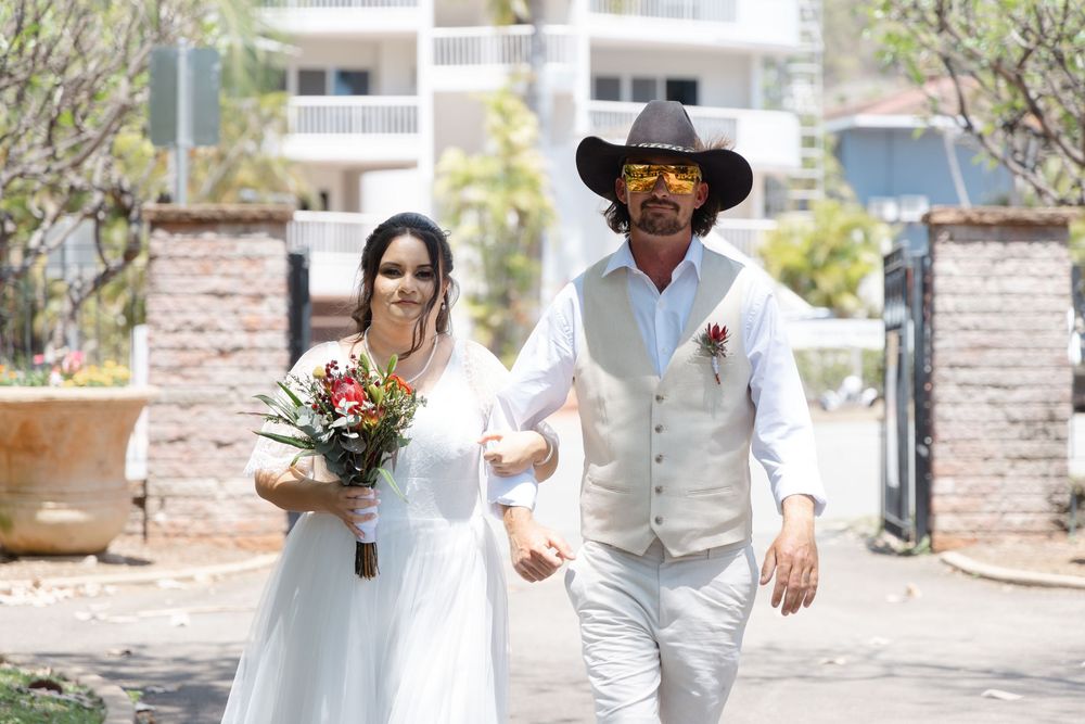A couple in matching white outfits walk together outdoors near stone pillars and greenery.