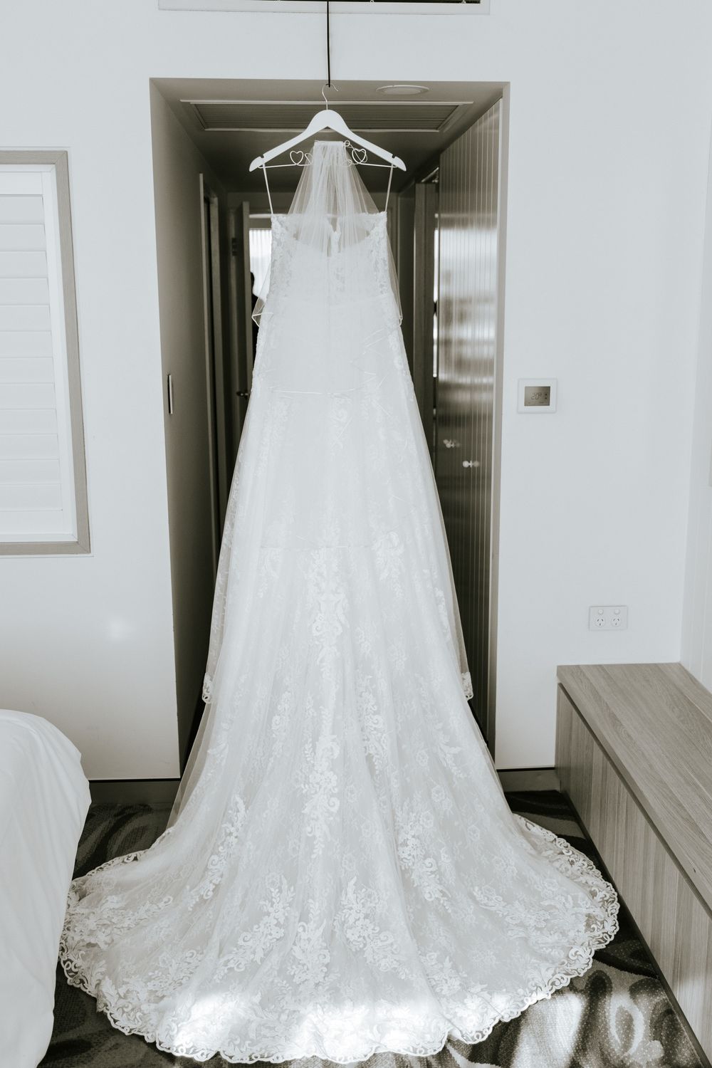 Elegant white lace wedding dress with long train displayed hanging in a hotel room hallway.
