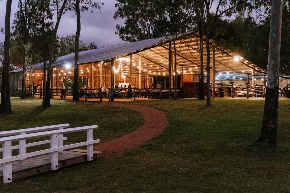 Rustic barn wedding venue illuminated at twilight with white fencing and trees in foreground.