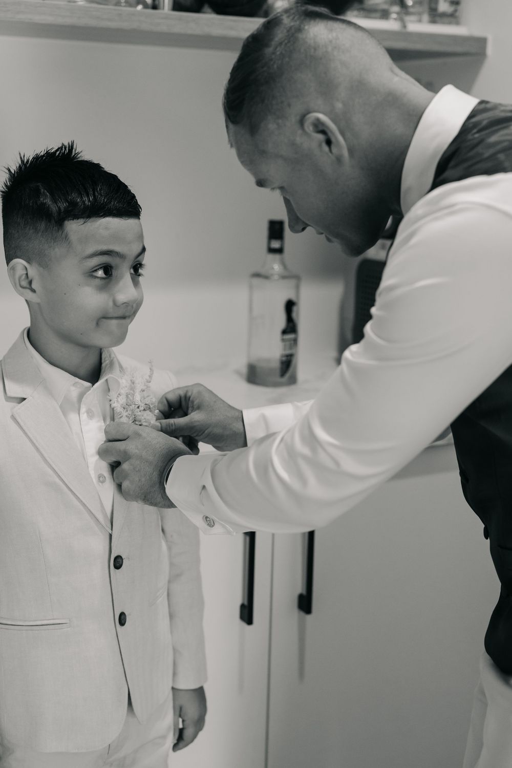 Black and white photo of a man in a vest pinning a boutonniere on a young boy in a white suit at a wedding.
