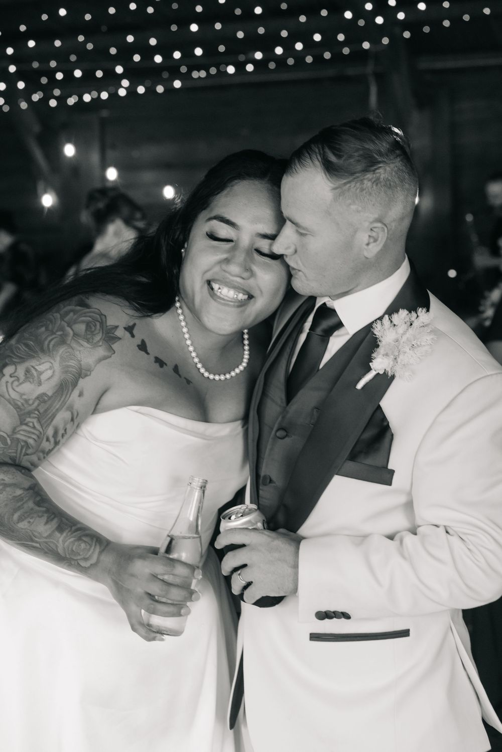 Black and white photo of a smiling bride with tattoos and pearl necklace being kissed by groom in white suit at wedding.