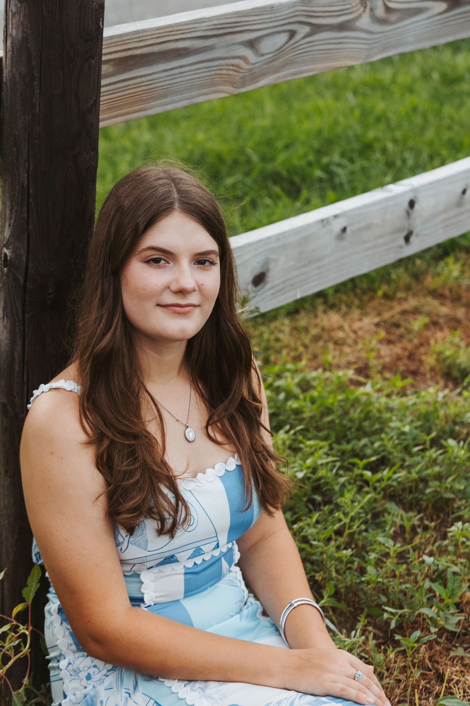 Someone in a light blue dress sits on a wooden fence in a grassy outdoor setting.