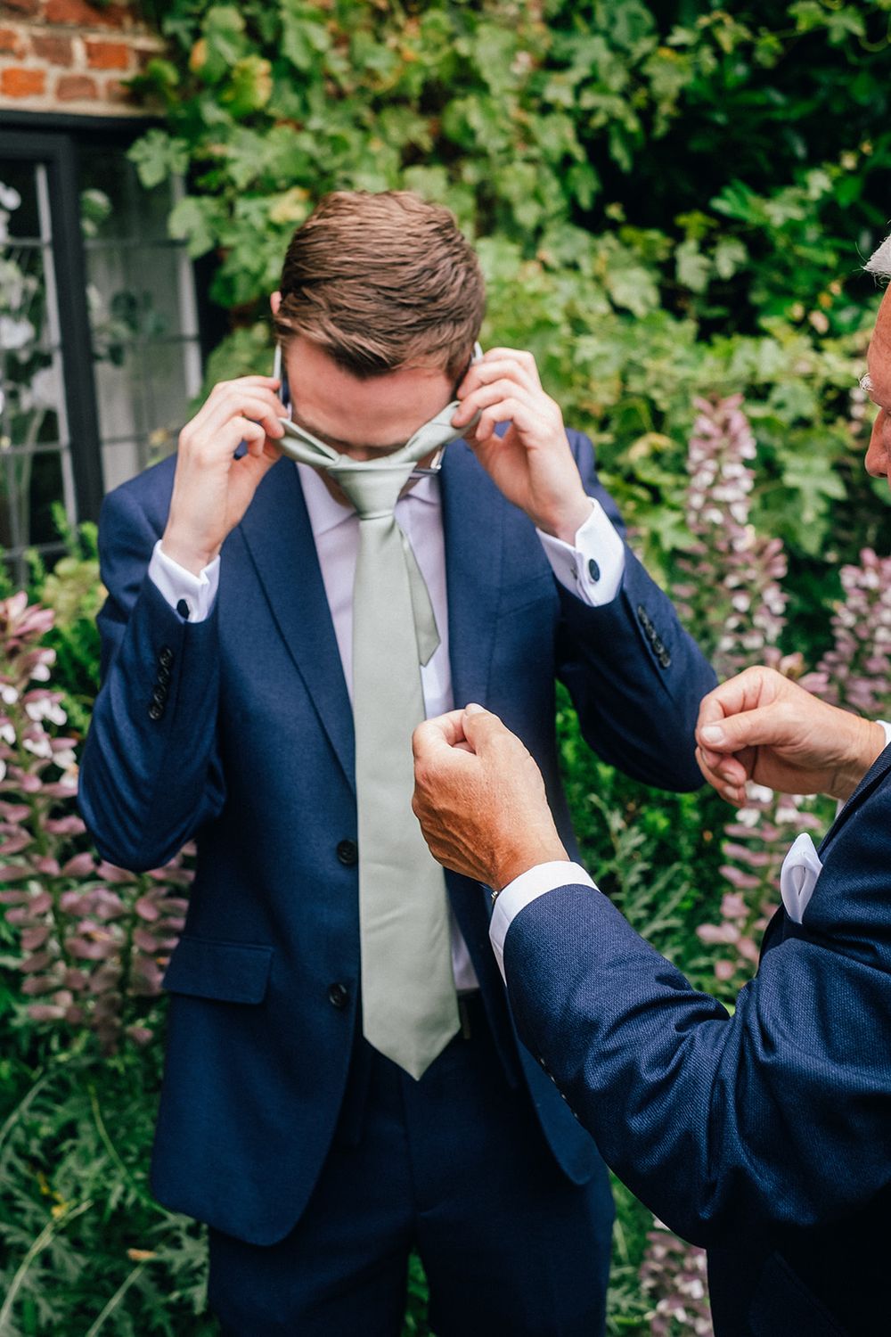 A person in a navy suit and light tie adjusts their attire while standing in front of greenery and brick wall.