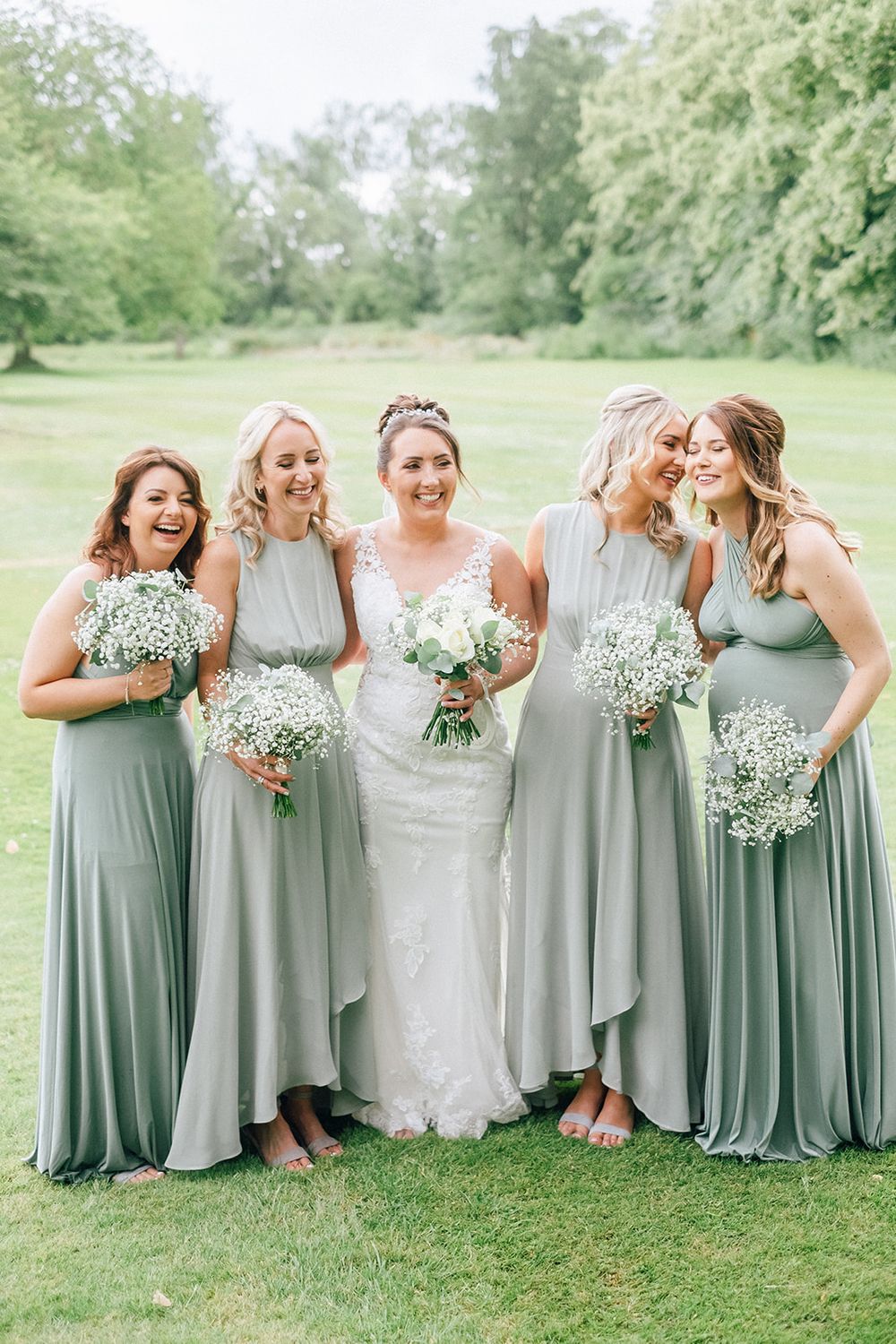 Wedding party in sage green dresses poses on grassy lawn while sharing candid laughs together.