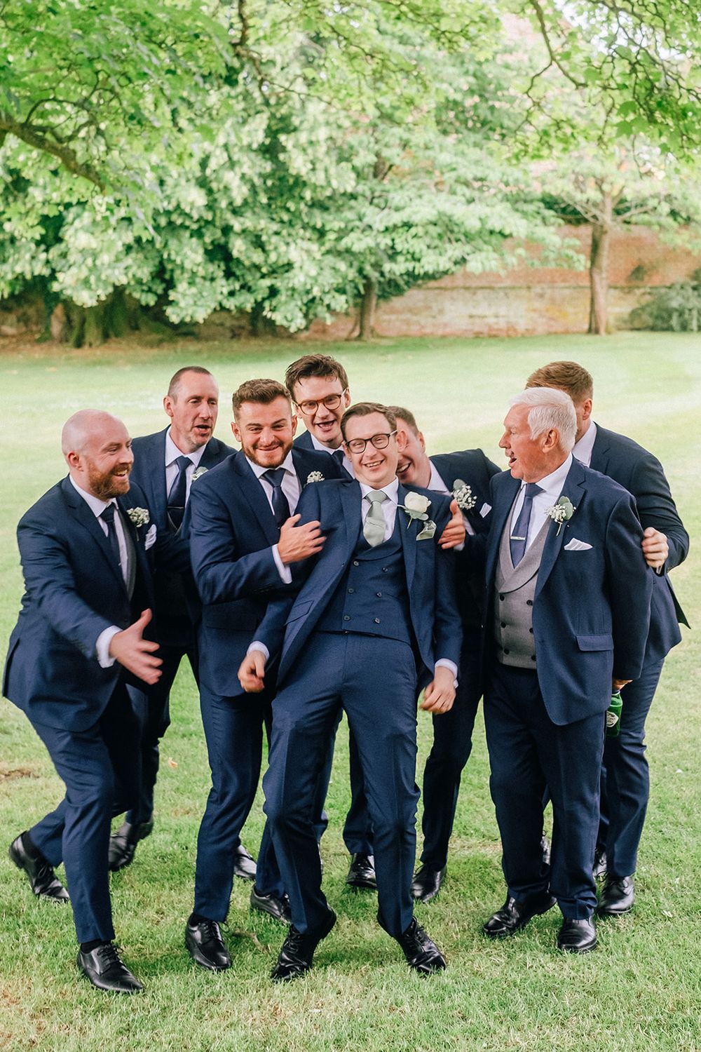 Groomsmen in navy suits share a candid laughing moment while posing on grass during outdoor wedding photos.