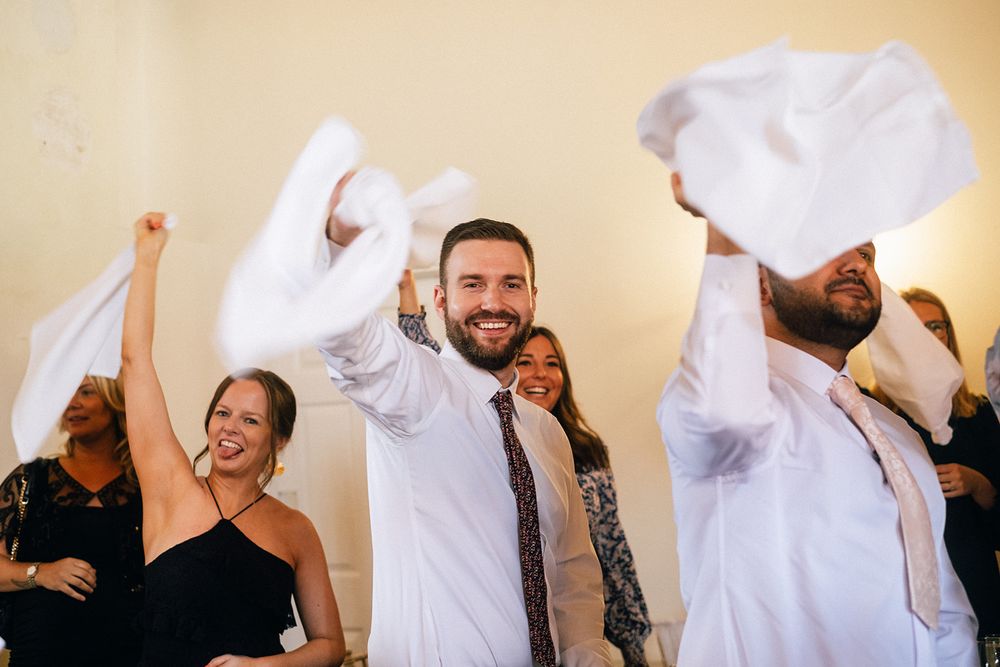 Wedding guests dance joyfully together at a celebration, waving white napkins in the air during a festive moment.