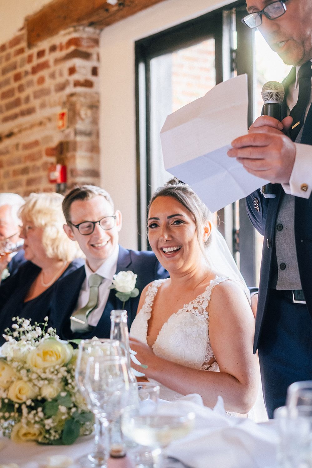 Wedding reception speech moment at rustic brick venue with floral centerpieces and table settings.